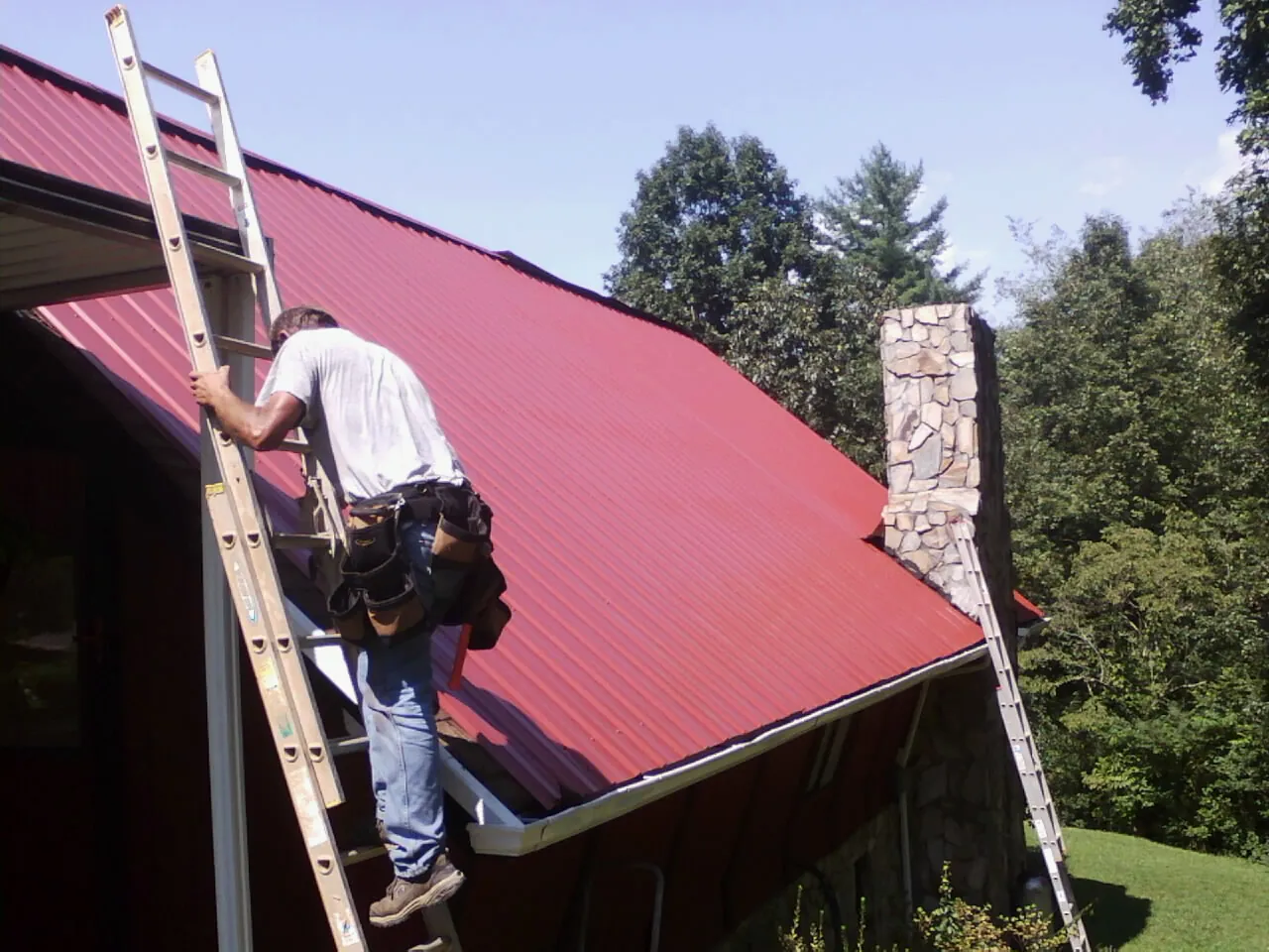 Roofer on a ladder beside a red metal roof and stone chimney — Soffit & Fascia Repair by Roofing Brothers in Morgan Hill